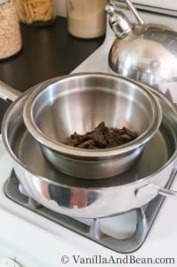 A stainless bowl with chopped chocolates is placed in a deep sauté pan with water heated on a burner.