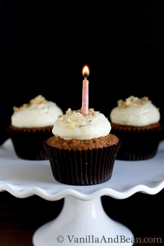 Three carrot cupcakes with icing on a cake stand. The middle cupcake has a small candle lit.