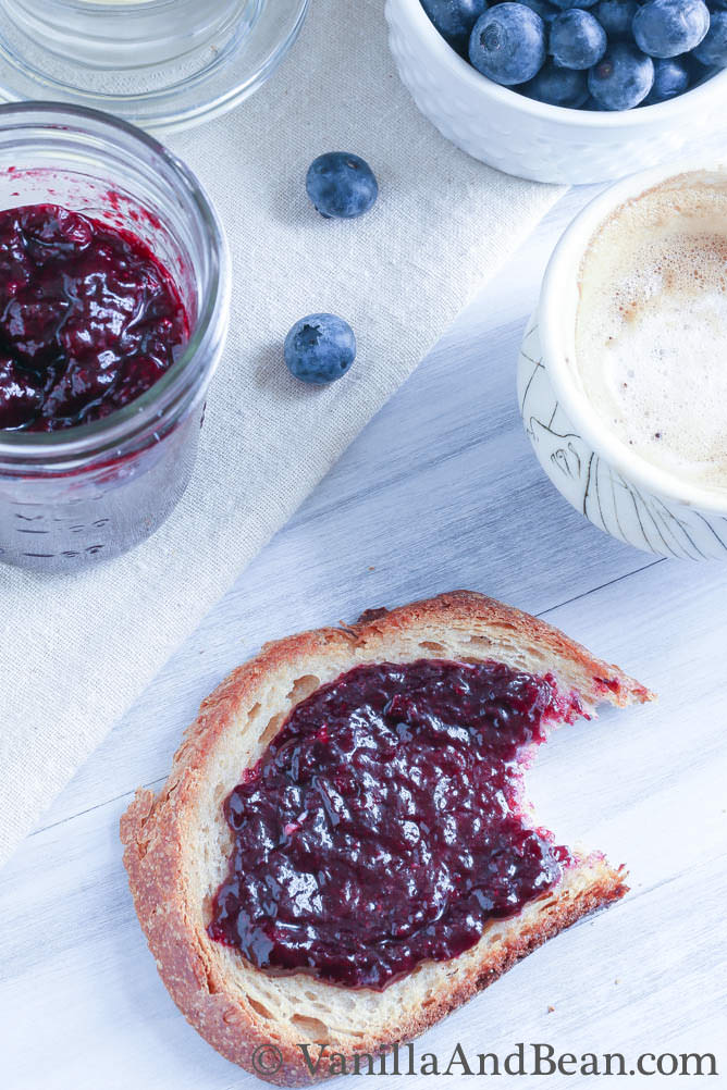 Blueberry jam in a small jar and some of the jam spread on a toasted bread.