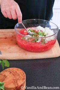 Sliced white onions added to the tomato mixture in a large glass bowl with a spoon