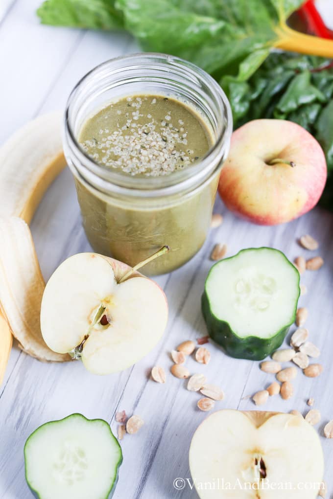 Green smoothie in a small mason jar surrounded by apples. cucumber, chard, and seeds.