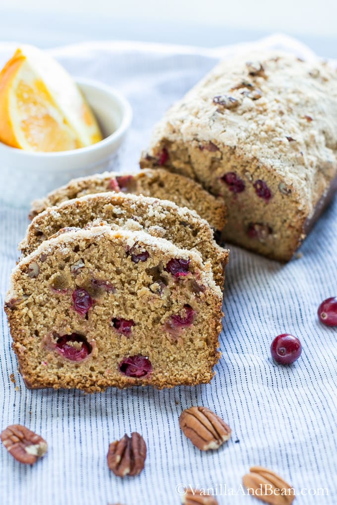 A loaf and three slices of the Cranberry Orange Bread with Pecans on a kitchen towel. A couple of orange wedges in a small bowl, pecans, and cranberries around it.