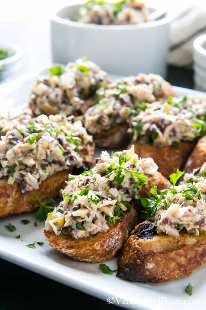 Olive Artichoke Crostini on a plate garnished with parsley and a small bowl of the artichoke mixture behind it.