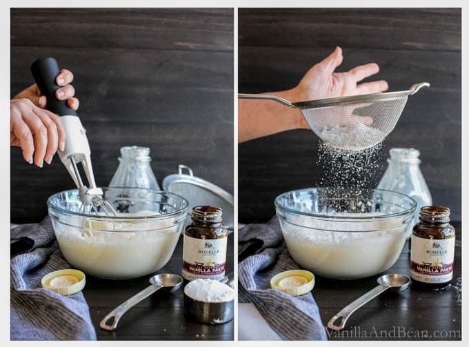 Heavy cream and vanilla bean paste in a large glass bowl mixed with a hand blender. Powdered sugar is sifted over the cream mixture that has thickened.