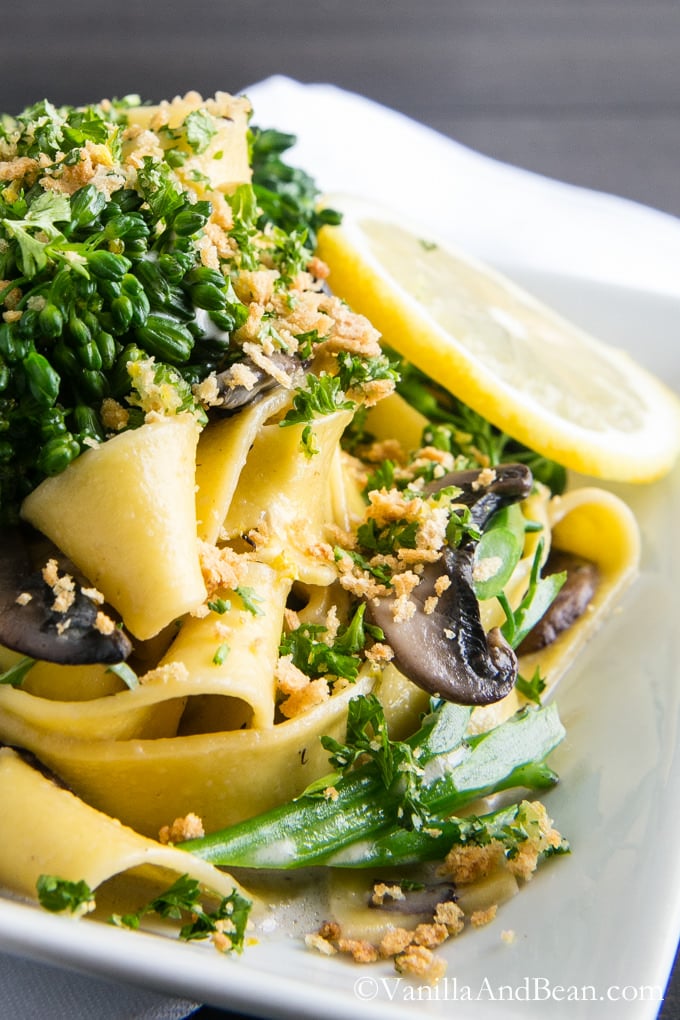 A plate of Pappardelle with Broccolini and Crunchy Gremolata and a slice of lemon.