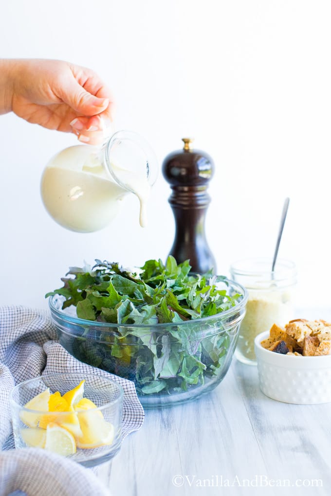 Caesar Dressing poured into a bowl of kale surrounded by a small bowl of croutons, a small bowl of lemon wedges and a glass of Parmesan mixture.