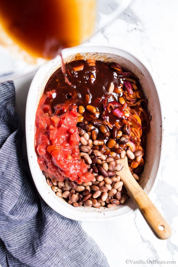 Mixing beans with carrots and other ingredients with a wooden spoon and adding the BBQ sauce to the beans. 