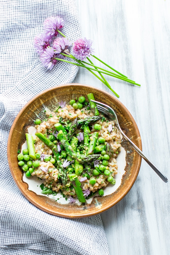 Creamy Farro with Pesto Asparagus and Peas in a flat bowl with a fork garnished with parsley, chives and chive flowers. 
