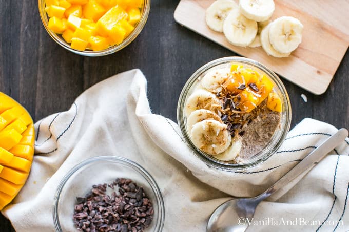A glass container of Creamy Coconut Porridge surrounded by cubed mangoes, sliced bananas and cocoa nibs.