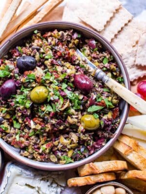 Olive tapenade recipe on a snack board with grapes, almonds, crostini, cheese and breadsticks.