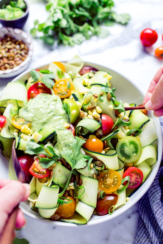 Tossing Raw Zucchini Ribbons Salad with corn and tomatoes in a bowl with avocado dressing.