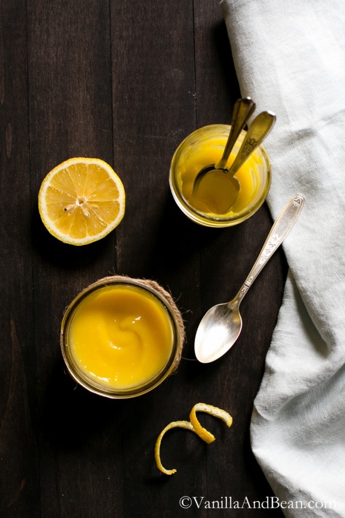 Lemon curd in jars with spoons and a half lemon on a table. 