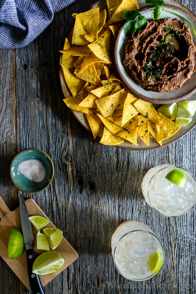 Chipotle Black Bean Dip on a table shared on a plate with tortilla chips, drinks with lime and salt. 