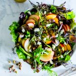 A big bowl of greens, pistachios, goat cheese and nectarines with dressing on the side ready to be shared.