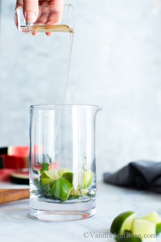 Pouring simple syrup into a mixing glass with mint and lime. 