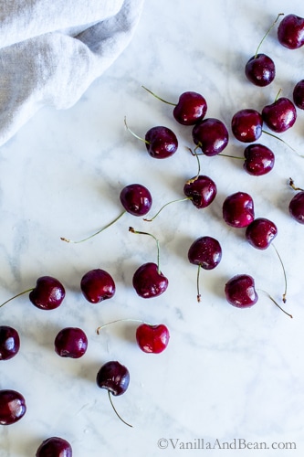Bing cherries lined up on a marble table with a tea towel off to the side. 
