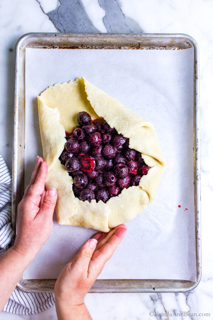 Cherry galette on a baking sheet with hands folding the pastry.