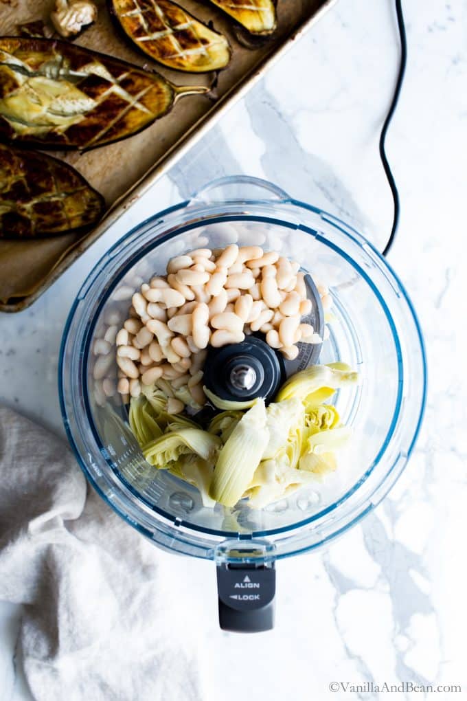 White beans, and artichokes in a food processor bowl with roasted eggplant standing by. 