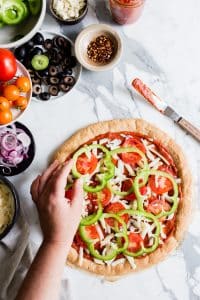 A pizza sitting on top of a marble table as person adds bell peppers to the top.