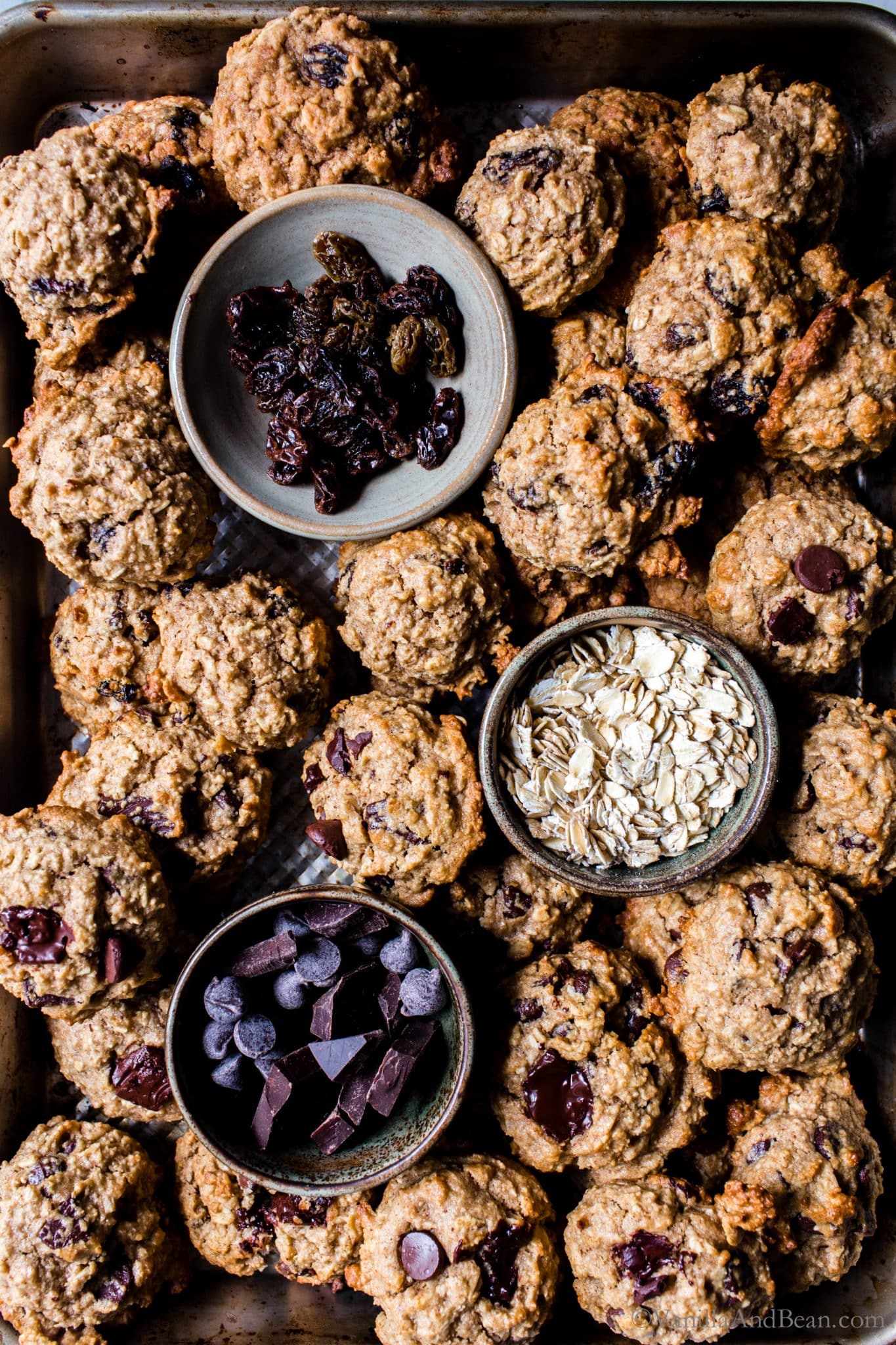 Baked Healthier Oatmeal Cookies on a pan with a bowl each of oats, raisins and chocolate chips. 