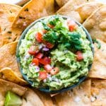 Creamy Guacamole Recipe in a bowl garnished with tomatoes and cilantro surrounded by tortilla chips.