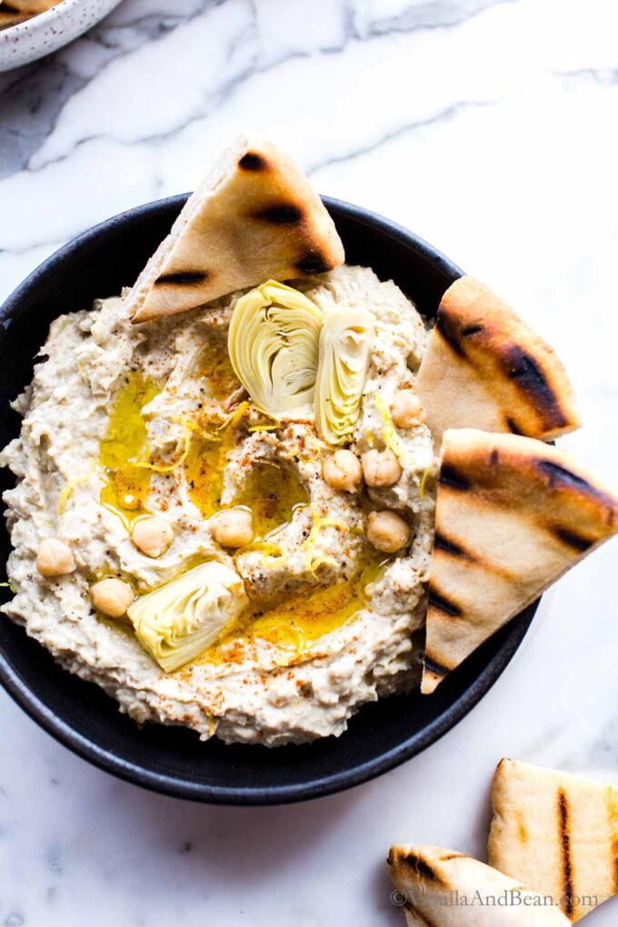 Close up of artichoke hummus dip in a bowl garnished with artichoke, chickpeas, olive oil and pita wedges. .