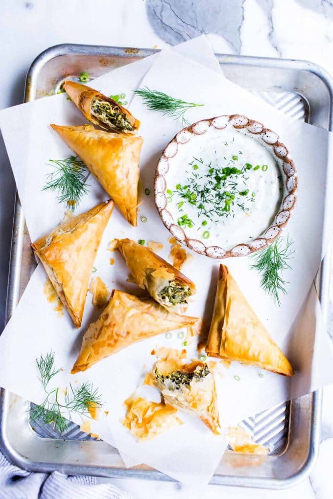 Spinach and feta parcels on a pan garnished with dill with a yogurt dip on the side.