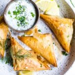 Spanakopita triangles on a plate garnished with dill with a yogurt dip on the side.