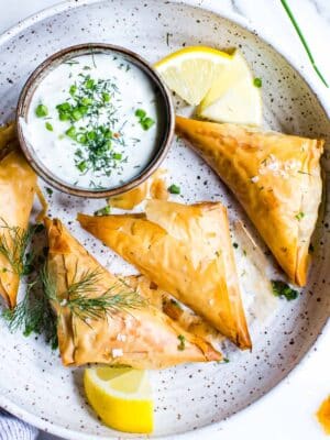 Spanakopita triangles on a plate garnished with dill with a yogurt dip on the side.