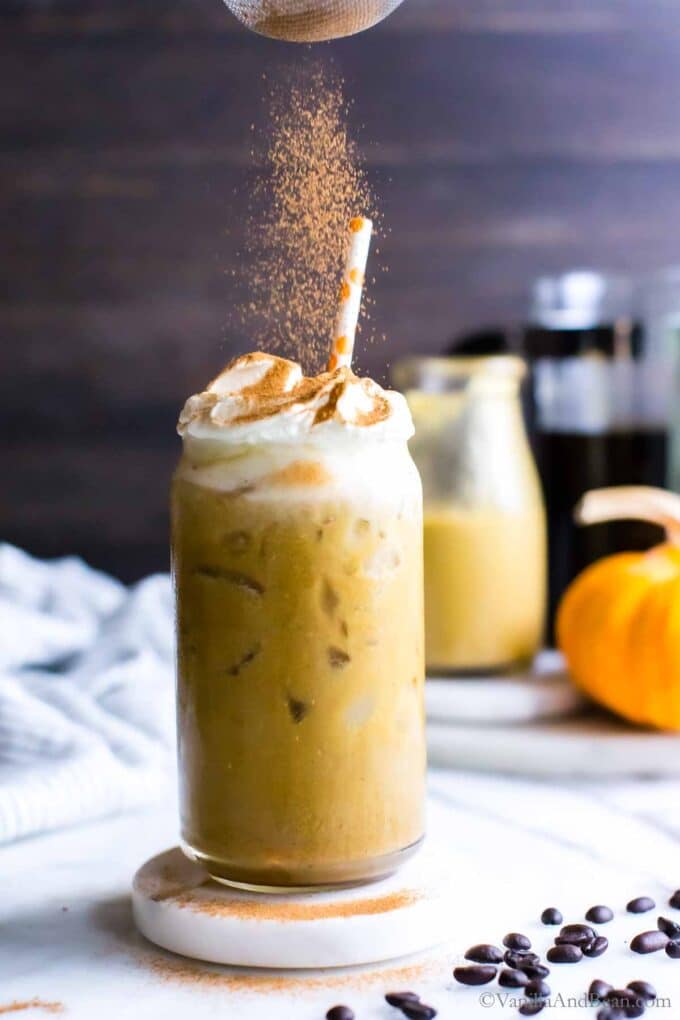 Pumpkin pie spice being sprinkled over the whipped cream in a iced pumpkin latte. 
