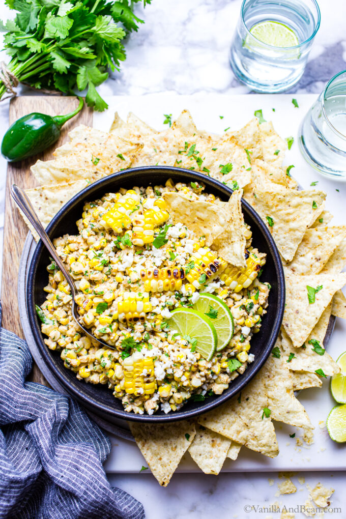 Elotes dip in a black bowl surrounded with tortilla chips, garnished with lime wedges, tortilla chips and cilantro.