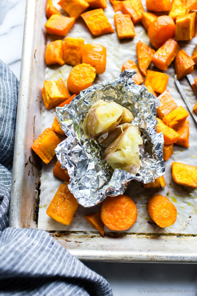 Roasted garlic in foil surrounded with roasted sweet potatoes and carrots.