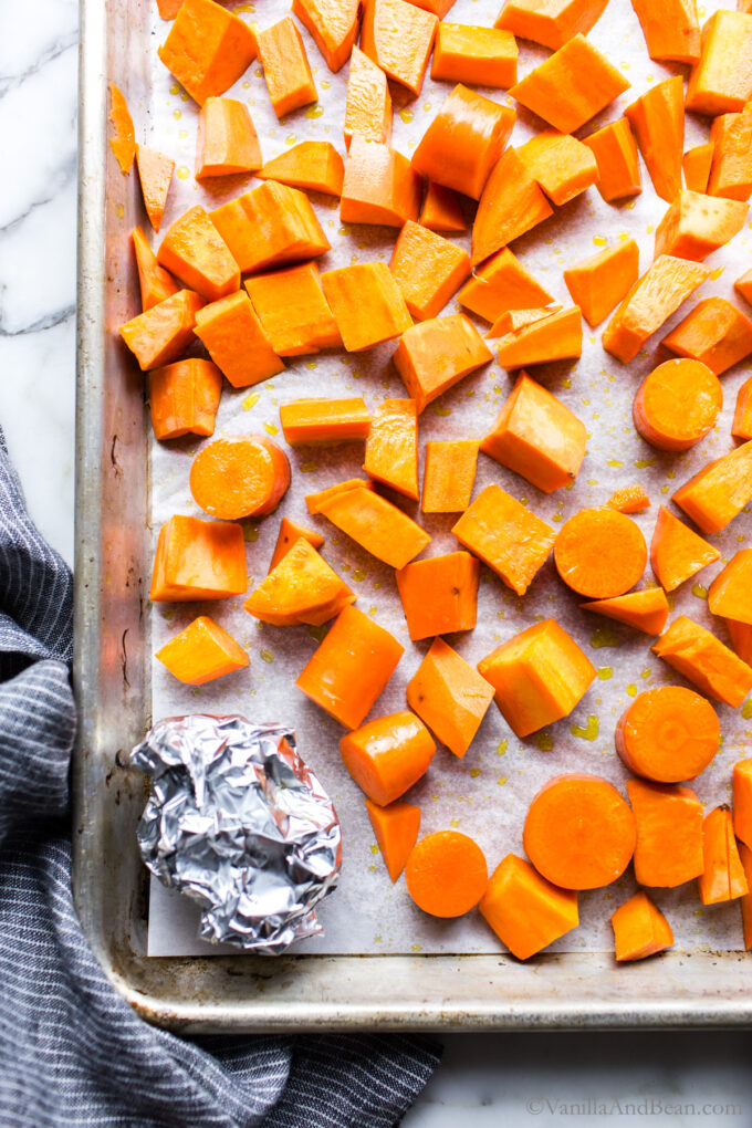 Sliced sweet potatoes and carrots on a sheet pan with a foil ball of garlic.