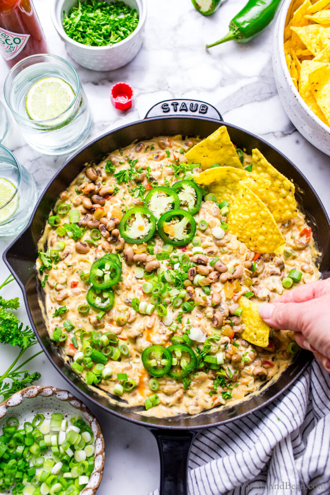 Creamy and warm Black Eyed Pea Dip with Cream Cheese in a skillet garnished with fresh jalapenos, green onions, parsley and tortilla chips with someones hand digging in. 
