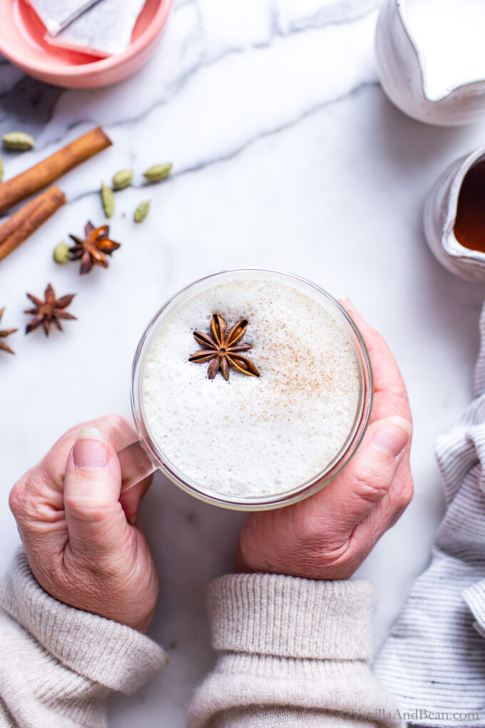 Hands holding hot chai tea latte in a mug with frothed milk and a star anise floating on top.