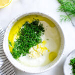 Yogurt and Dill Sauce in a bowl with ingredients showing on top: dill, garlic, lemon, olive oil.