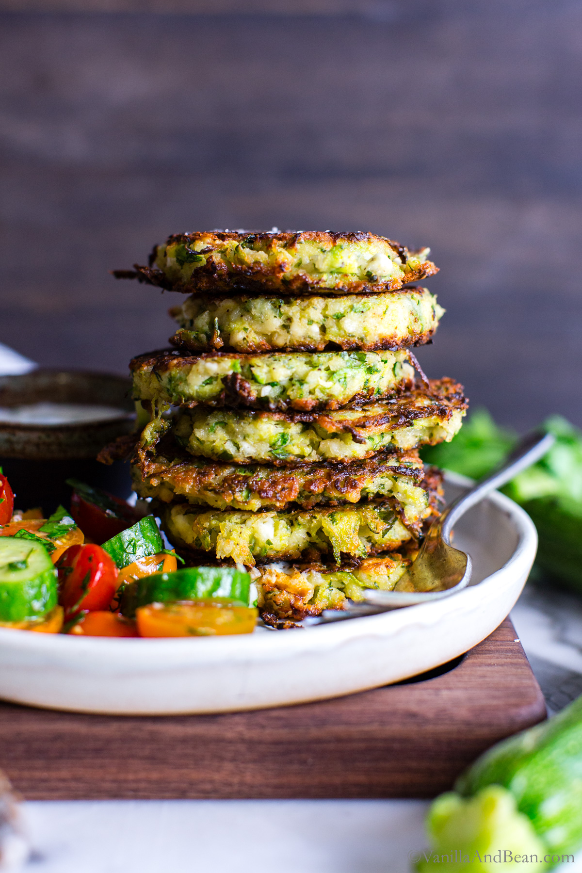 Zucchini and feta fritters stacked stacked tall on a plate with a simple tomato salad.