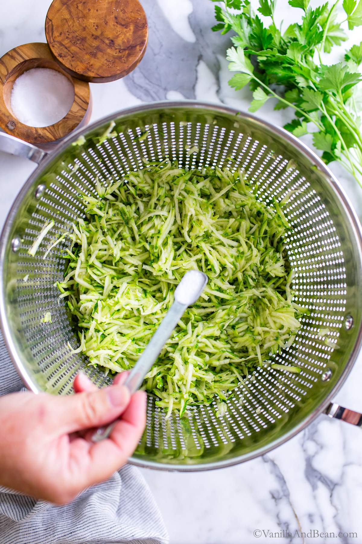 Grated zucchini in a colander with salt being sprinkled in.